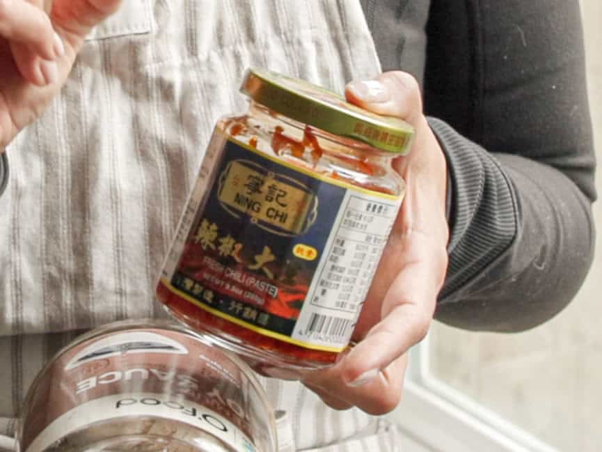 A person wearing an apron holds a jar of Ning Chi brand chili paste in one hand and another jar with an obscured label in the other.